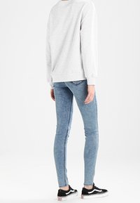 Světle šedá mikina, přiléhavé modré džíny a černé tenisky. Džíny mají opranou texturu a viditelné švy podél stran.