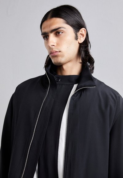Schwarze Zip-Jacke mit hohem Kragen, glattem Stoff, minimalistischem Design und einem Knopfdetails am Hals.