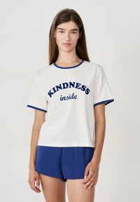 Joven mujer vestida con una camiseta blanca con ribete azul y el texto "KINDNESS inside", combinada con pantalones cortos azules, de pie frente a un fondo sencillo.