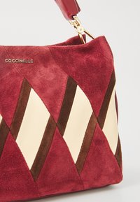 Red suede handbag featuring diamond-patterned design with cream and brown accents. Gold hardware and a burgundy strap complete the look.