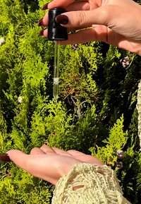 Dłoń z ciemnym lakierem na paznokciach dozująca płyn z zakraplacza do otwartej dłoni na tle zielonych liści.