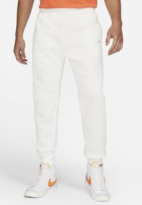 White sweatpants with an elastic waistband and a small logo on the left thigh, paired with white sneakers featuring orange accents.