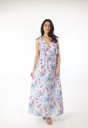 Mujer con un vestido maxi azul de flores sin mangas, con tirantes anudados en los hombros, de pie descalza sobre un fondo blanco.