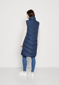 Ναυτικό γιλέκο πούφερ με ψηλό γιακά και σχέδιο βελόνας. Φορεμένο πάνω από ένα ανοιχτόχρωμο μπλουζάκι, σε συνδυασμό με μπλε τζιν και λευκά αθλητικά παπούτσια.