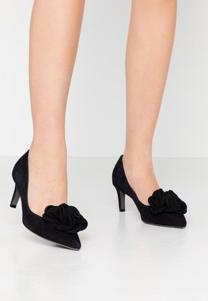 Czarne zamszowe szpilki z wydłużonym noskiem i zakrzywionym designem, z charakterystycznym kwiatowym akcentem na nosku. Wysokość obcasa 3 cale.