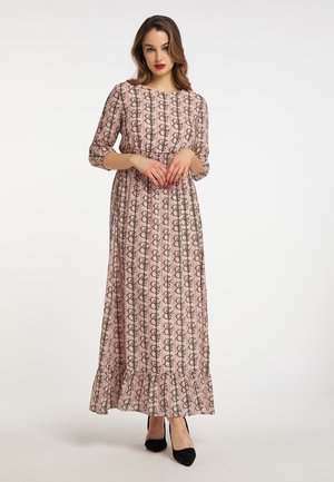 Vrouw die een lange roze jurk met patroon en 3/4 mouwen draagt en zwarte hoge hakken staat tegen een effen witte achtergrond.