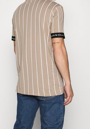 Mand iført beige kortærmet skjorte med tynde lodrette hvide striber og sorte ærmekanter, kombineret med blå jeans, set bagfra.