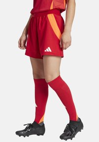 Rode sportieve shorts met oranje accenten, voorzien van een elastische tailleband, gecombineerd met rode kniehoog sokken en zwarte voetbalschoenen.
