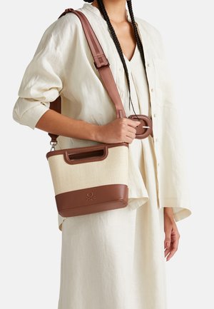 Nainen, jolla on punotut hiukset ja joka on pukeutunut kermanväriseen asuun, pitää käsissään beigeä ja ruskeaa crossbody-laukkua, jossa on leveä säädettävä hihna ja solkikuvio.