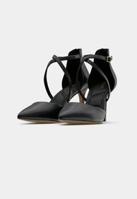 Svarte spisse pumps med høye hæler, kryssede ankelsnorer og gullspenne detaljer mot hvit bakgrunn.