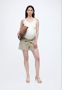 Femme enceinte en débardeur blanc, short beige en dentelle, tenant un sac marron, portant des sandales à talons beiges, debout sur fond blanc.