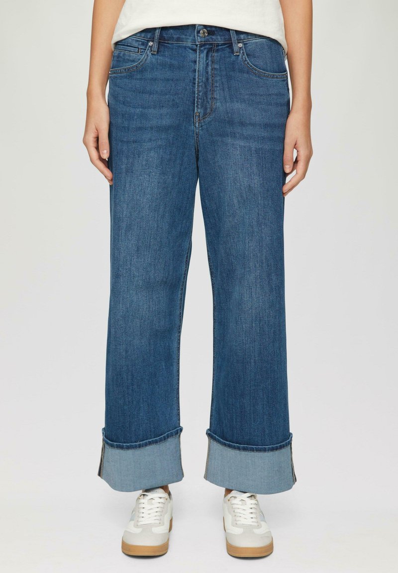 Brede blauwe denim jeans met een opgezoomde zoom en een lichte vervaging, met vijf zakken en een knoopsluiting bij de taille. Gecombineerd met witte sneakers.
