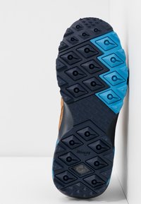 Semelle d'une chaussure de randonnée avec un motif de crampons hexagonaux noirs et bleus et une adhérence texturée, étiquetée "Hexalite", inclinée contre une surface blanche.