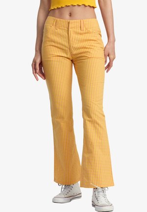 Mujer con pantalones acampanados a cuadros en amarillo y naranja con bolsillos delanteros y zapatillas altas blancas sobre fondo blanco.