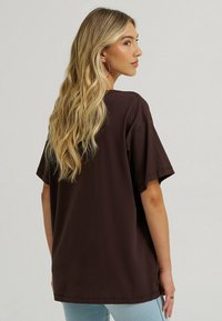 Brązowy, oversize t-shirt z krótkimi rękawami i okrągłym dekoltem. Tkanina wydaje się miękka o gładkiej fakturze. Na sobie ma jasne dżinsy.