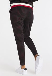 Pantalones negros con rayas rojas, ajuste tapered, cinturilla elástica con una franja roja y blanca, combinados con zapatillas blancas.