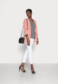 Blazer rosa claro sobre uma blusa padrão em preto e branco, calças brancas e sapatos de salto pretos com tiras. Uma pequena mala de ombro preta completa o visual.