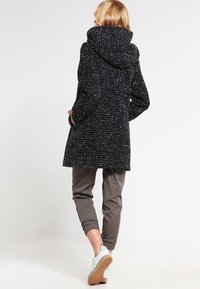 Cappotto a pois bianco e nero con cappuccio, design fino al ginocchio, tessuto testurizzato. Abbinato a pantaloni grigi e sneakers bianche.