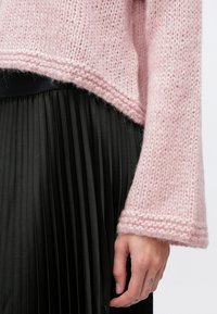 Ροζ πλεκτό πουλόβερ με άνετη εφαρμογή και ριμπ μανσέτες, συνδυασμένο με μαύρη πλισέ φούστα. Το ύφασμα φαίνεται μαλακό και με υφή.