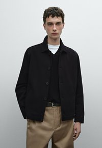 Giacca nera a bottoni realizzata in un materiale morbido, indossata sopra un maglione a V scuro, abbinata a pantaloni beige chiaro e una cintura.