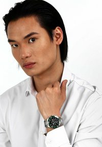 Chronograph-Uhr mit schwarzem Ziffernblatt, grünem Lünette und silbernem Metallarmband. Am Handgelenk getragen, Modell in einem weißen Hemd mit Kragen.