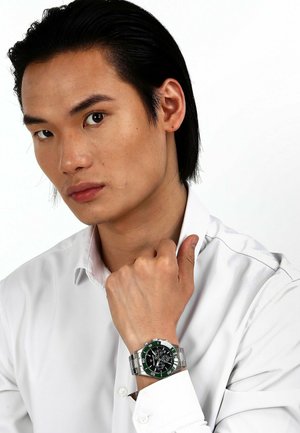 Chronografklocka med svart urtavla, grön bezel och silvermetallarmband. Buren på ett handled, modellen i en vit skjorta med krage.