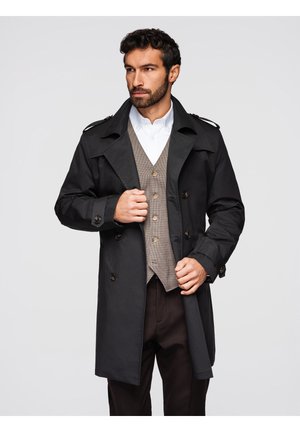 Mann trägt schwarzen Trenchcoat über einer braun karierten Weste und weißem Hemd, richtet seine Mantelknöpfe, vor einem schlichten grauen Hintergrund.