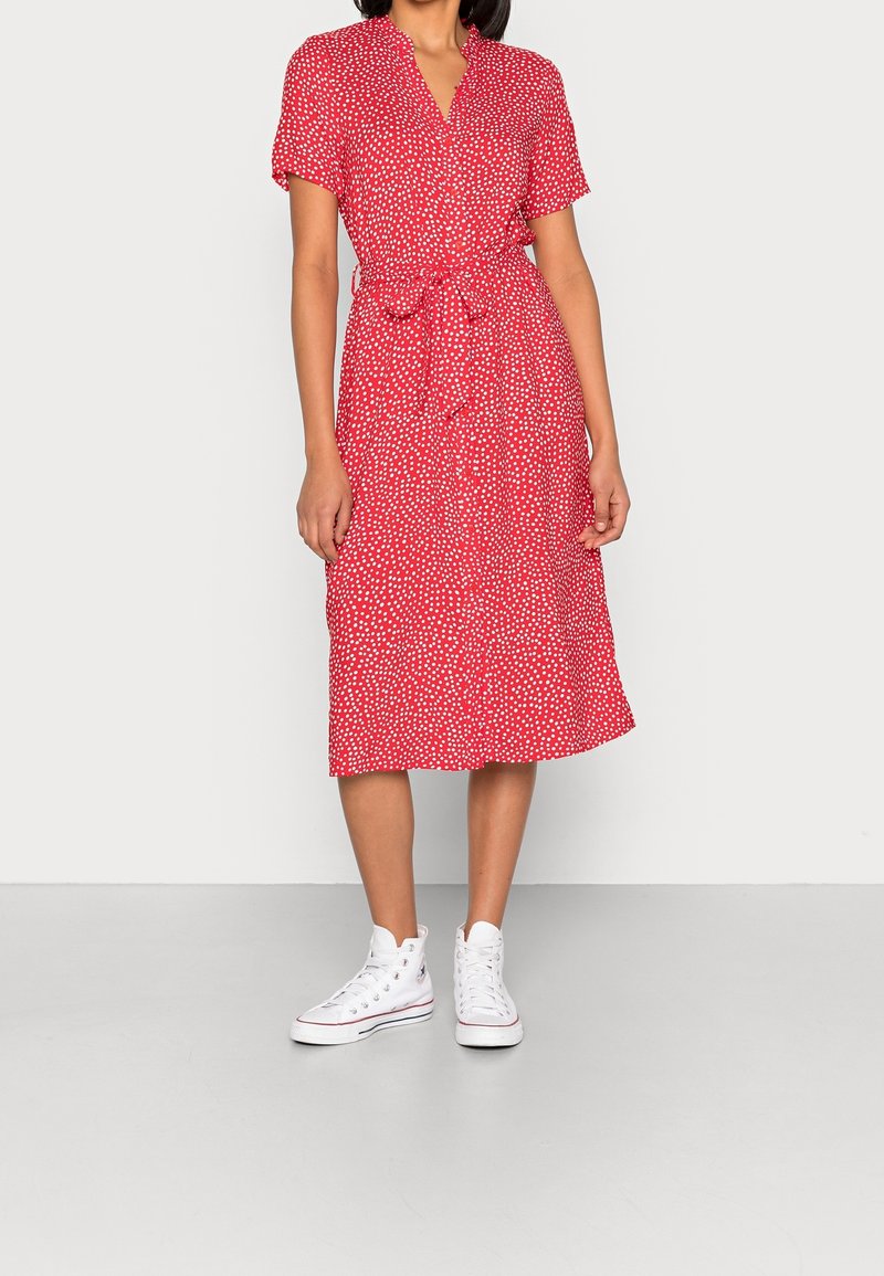 Femme portant une robe midi rouge à manches courtes avec des pois blancs, taille cintrée et baskets montantes blanches, debout sur un sol gris.