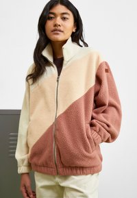 Chaqueta de forro polar con cierre delantero, diseño a bloques de color en crema, beige y óxido, con una superficie texturizada y puños elásticos.