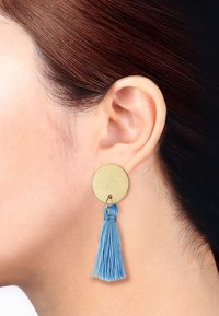 Runde Ohrringe aus goldfarbigem Metall mit einer blauen Quaste. Die strukturierte Oberfläche der Scheibe steht im Kontrast zu den glatten Quastensträngen.