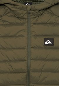 Olivgrüne gesteppte Jacke mit zentralem Reißverschluss und kleinem schwarz-weißen Logopatch auf der linken Brust.