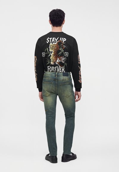 Homme portant un jean bleu délavé et une chemise noire à manches longues avec un motif tigre et le texte "Stay Up Forever" dans le dos.
