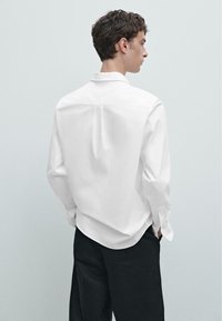 Camicia bianca a maniche lunghe con una texture morbida, plissé posteriore e colletto classico. Indossata con pantaloni neri, mostrata da un'angolazione laterale.