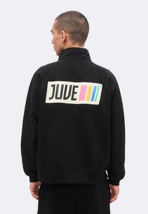 Muškarac u crnoj jakni s visokim ovratnikom, s natpisom "JUVE" i ružičastim, žutim i plavim okomitim prugama na leđima.