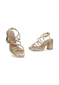 Sandalias de tacón alto con tiras en dorado metálico. Cuentan con un tacón cuadrado, punta abierta y múltiples tiras entrecruzadas a lo largo del pie.