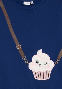 Tmavomodrá mikina s aplikáciou cupcakes s usmievavou tvárou, hnedými popruhmi so zlatými prvkami a mäkkým, textúrovaným povrchom.