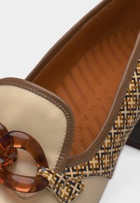 Γυναικείο ταμπά και μπεζ μοκασίνι με διακοσμητικές πλευρές με σχέδια και μεγάλο καφέ διακοσμητικό αλυσίδα στο μπροστινό μέρος.