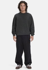 Dunkelgrauer gesprenkelter Pullover mit rundem Halsausschnitt, getragen mit lockeren schwarzen Hosen und braunen Turnschuhen, vor einem schlichten Hintergrund stehend.