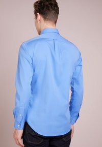Světle modrá košile s dlouhým rukávem z hladké látky, s špičatým límcem a zaobleným lemem. Knoflíkové detaily na manžetách.