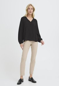 Kvinde står iført en sort V-hals trøje med lange ærmer, beige slim-fit bukser og sorte loafers foran en helt lys baggrund.