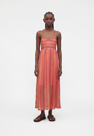 Kvinne som står vendt mot kamera iført en ermeløs, ankel-lang flerfarget stripete kjole med tynne stropper og brune sandaler.