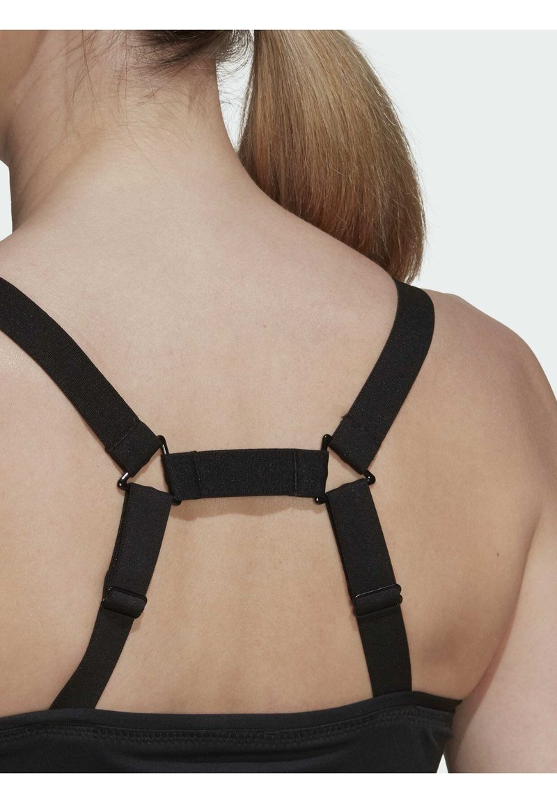 Rückansicht einer Person mit heller Haut und roten Haaren, die ein schwarzes Top mit verstellbaren Trägern trägt, bei dem ein zentraler horizontaler Riemen die Schulterträger verbindet.