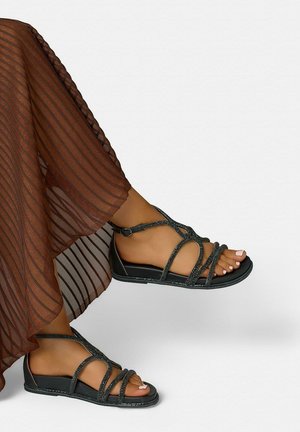 Opalone nogi w czarnych płaskich sandałach z paseczkami i przezroczysta brązowa plisowana spódnica na jasnym tle.