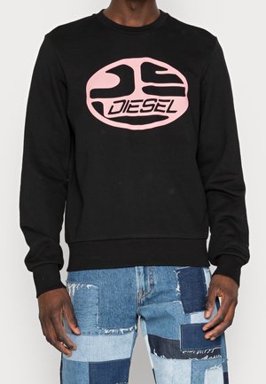 Personne portant un sweat-shirt noir avec un logo Diesel rose et un jean patchwork bleu aux différentes nuances de denim et aux bords effilochés.
