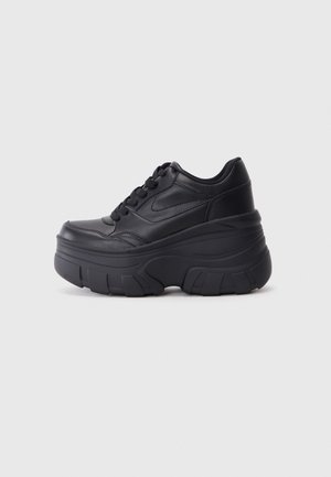 Zwarte leren plateau sneaker met dikke getextureerde zool en veterontwerp, zijaanzicht op een effen lichtgrijze achtergrond.