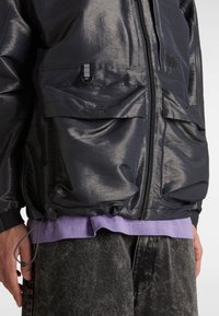 Veste noire zippée avec grandes poches avant et logo Vans, portée sur une chemise violette et un pantalon gris foncé.