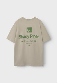 Beige katoenen t-shirt met een groen grafisch ontwerp op de rug, met de tekst "Shady Pines Golf Club" en een golfbalicoon.