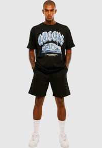 Black graphic t-shirt with "QUEENS" in bold, blue lettering and a car design. Paired with black shorts and white sneakers with crew socks.
