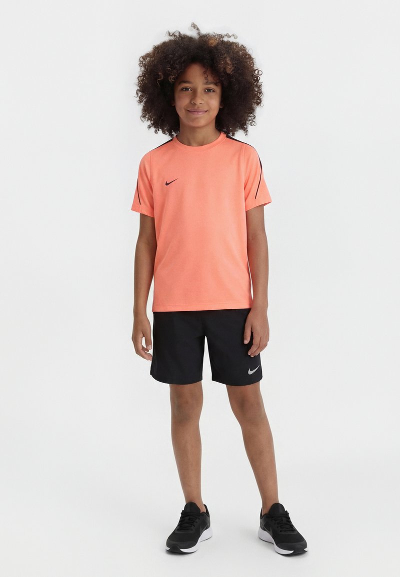 Παιδί με σγουρά μαλλιά που φορά ροζ κοραλλί μπλουζάκι Nike, μαύρο σορτς Nike και μαύρα αθλητικά παπούτσια, στέκεται μπροστά σε λευκό φόντο.