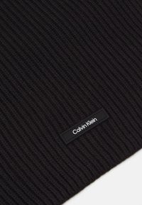 Beanie in maglia nera a coste con una superficie testurizzata e un'etichetta rettangolare nera con testo bianco "Calvin Klein".